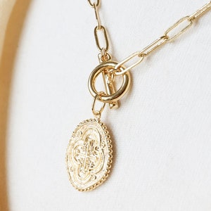 May include: A gold chain necklace with a large, oval-shaped pendant featuring a detailed, intricate design. The necklace has a toggle clasp.