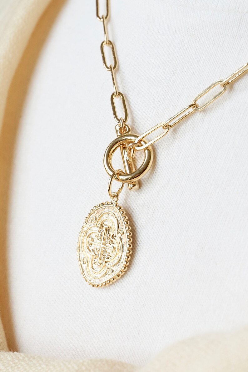 May include: A gold chain necklace with a large, oval-shaped pendant featuring a detailed, intricate design. The necklace has a toggle clasp.