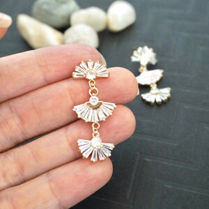 May include: Gold-toned, three-tiered earrings with a fan-like design. Each tier features a central clear stone surrounded by baguette-cut stones. The earrings are connected by delicate gold chains, creating a dangling effect.