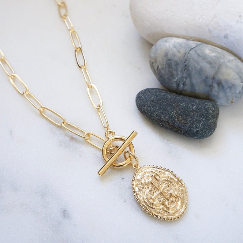 May include: A gold necklace featuring a toggle clasp and a large, oval pendant with an intricate design. The pendant is textured and has a raised, circular border.
