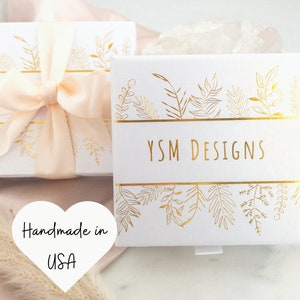 May include: Two white gift boxes with gold leaf designs and the text "YSM DESIGNS". One box is tied with a peach ribbon. The image also includes a heart-shaped graphic that says "Handmade in USA".