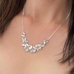 May include: A silver necklace with a delicate chain and a floral design. The flowers are arranged in a curved pattern and have a slightly textured finish.