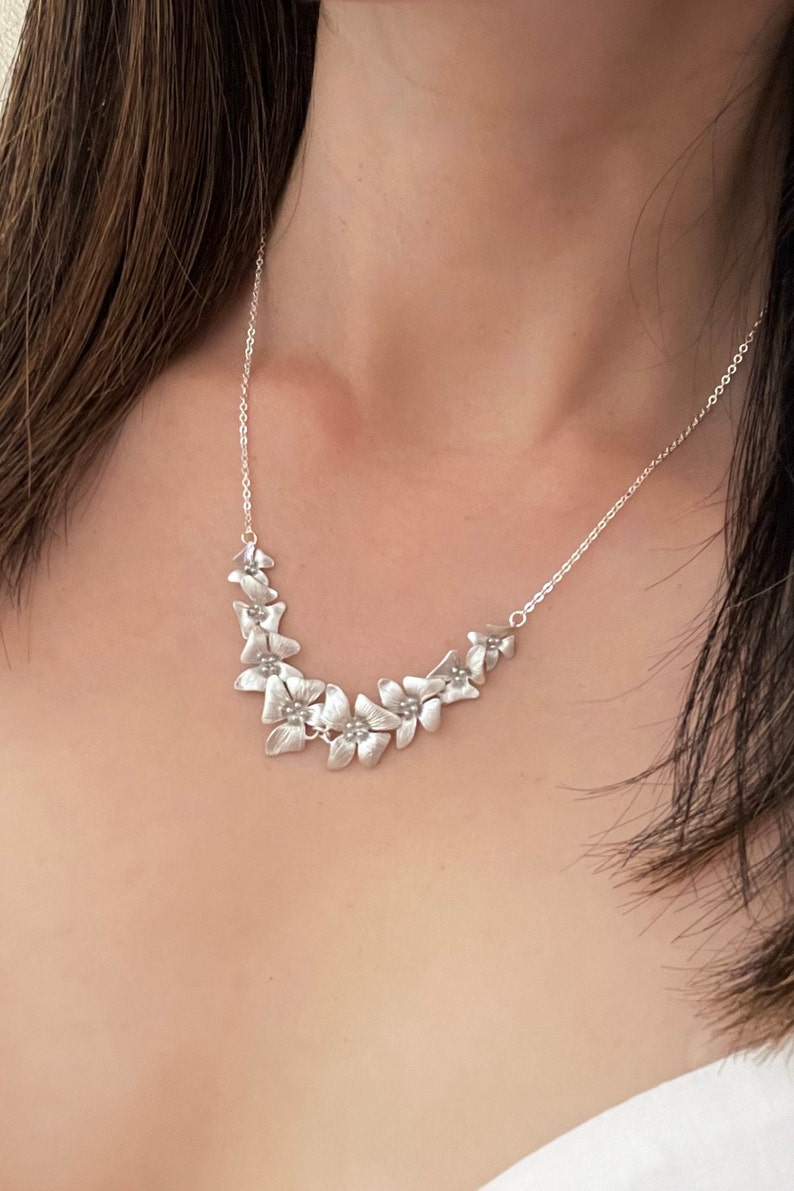 May include: A silver necklace with a delicate chain and a floral design. The flowers are arranged in a curved pattern and have a slightly textured finish.