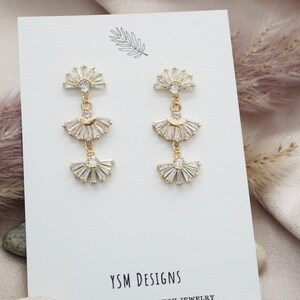 May include: Gold-toned drop earrings with three fan-shaped sections, each adorned with clear crystals. The earrings have a post-back closure and are displayed on a white card with the text "YSM DESIGNS". The design is elegant and suitable for special occasions.