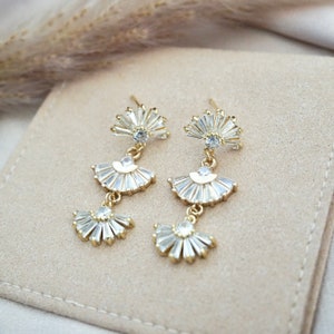 May include: A pair of gold-toned drop earrings with a fan-shaped design. Each earring features three tiers of clear, baguette-cut crystals, suspended from a small, round crystal. The earrings are displayed on a beige surface.