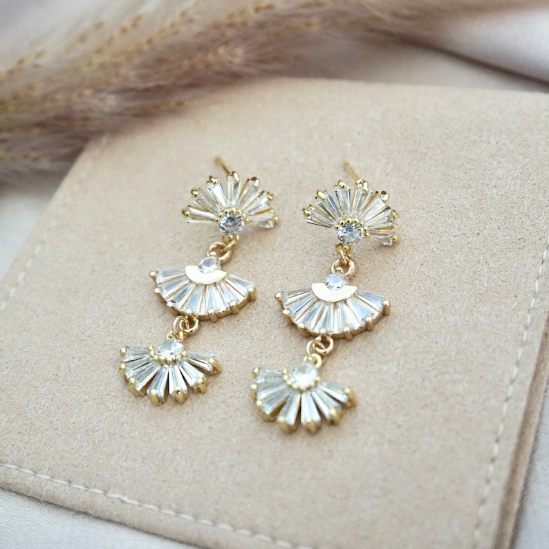 May include: A pair of gold-toned drop earrings with a fan-shaped design. Each earring features three tiers of clear, baguette-cut crystals, suspended from a small, round crystal. The earrings are displayed on a beige surface.