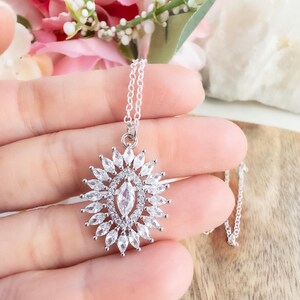 May include: A silver necklace with a pendant featuring a central marquise-cut clear stone surrounded by numerous smaller, clear, marquise-cut stones. The pendant is suspended from a delicate silver chain. The necklace is displayed against a soft background.