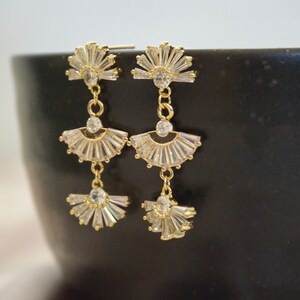 A pair of gold Art Deco earrings hanging on a black backdrop.  The earrings consist of 3 crystal studded fan shaped pieces connected together