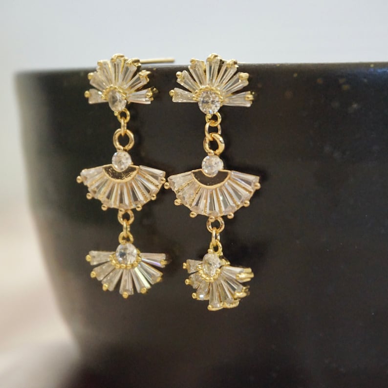 A pair of gold Art Deco earrings hanging on a black backdrop.  The earrings consist of 3 crystal studded fan shaped pieces connected together