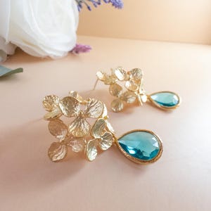Peut inclure: Boucles d'oreilles pendantes florales dorées avec un groupe d'éléments en forme de fleurs. Chaque boucle d'oreille présente une pierre précieuse bleu clair en forme de goutte. Les boucles d'oreilles sont posées sur un fond neutre et doux.