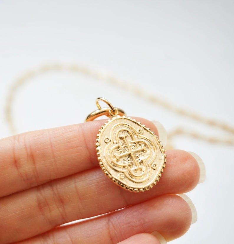 May include: Gold chain necklace with a large oval pendant featuring a detailed, embossed cross design.