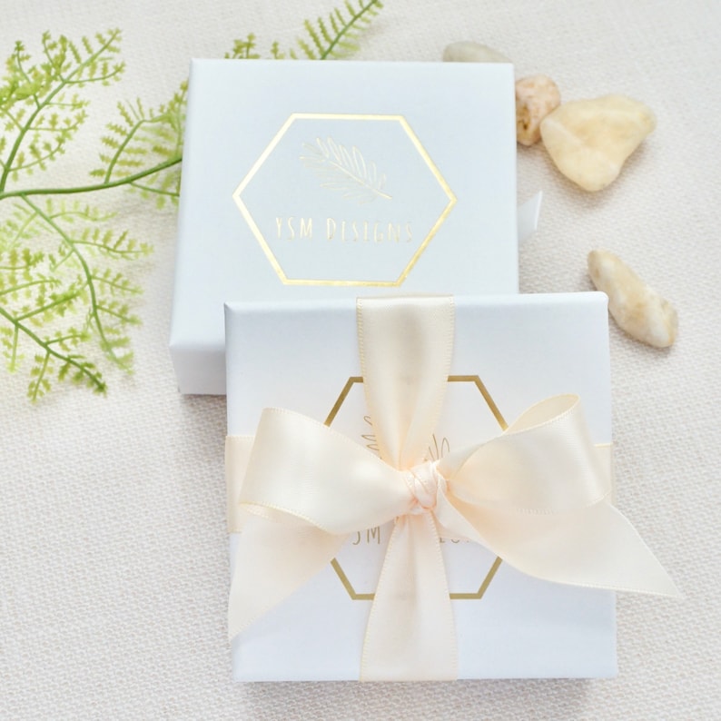 May include: Two white gift boxes with gold lettering. The top box has the text "YSM Designs" and the bottom box has a gold ribbon tied in a bow.