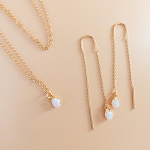 May include: Gold-toned jewelry set featuring a necklace and a pair of earrings. The necklace has a delicate chain with a teardrop-shaped, white gemstone pendant. The earrings are threader style, with a similar teardrop gemstone. The jewelry is displayed on a light beige surface.