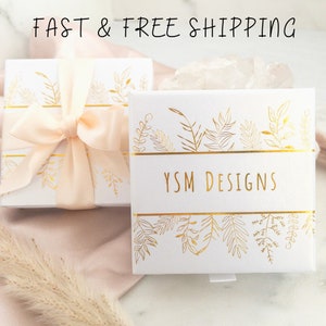K&ouml;nnte beinhalten: Eine wei&szlig;e Geschenkbox mit einem pfirsichfarbenen Band und goldenen Blumenmustern. Auf der Box steht "YSM DESIGNS" gedruckt.