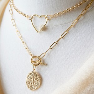 May include: Two gold necklaces layered on a white mannequin. The top necklace features a heart-shaped clasp with clear crystals. The bottom necklace has a toggle clasp and a round pendant with a cross design.