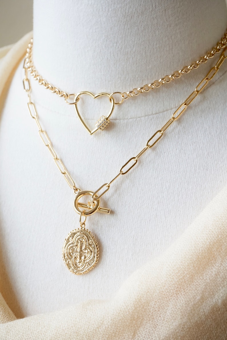 May include: Two gold necklaces layered on a white mannequin. The top necklace features a heart-shaped clasp with clear crystals. The bottom necklace has a toggle clasp and a round pendant with a cross design.