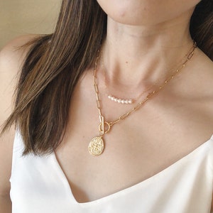 May include: A gold chain necklace with a large, ornate pendant and a smaller chain with a row of pearls. The necklace is layered and worn by a person with long brown hair.