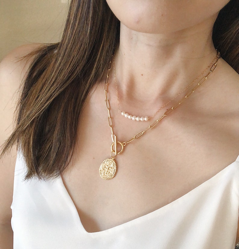 May include: A gold chain necklace with a large, ornate pendant and a smaller chain with a row of pearls. The necklace is layered and worn by a person with long brown hair.