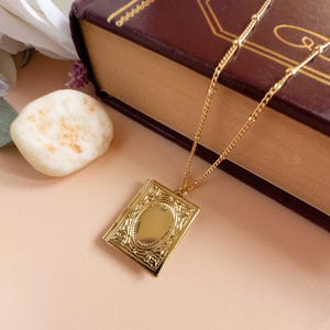 May include: A gold-toned metal locket pendant shaped like a small rectangular book with an embossed floral border and a central oval plate, suspended from a delicate gold-toned chain featuring small evenly spaced beads along the length of the chain.