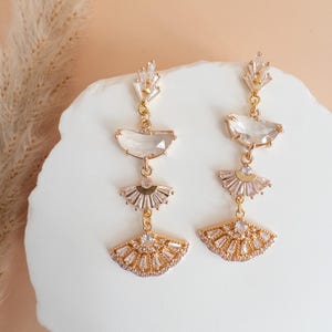 May include: A pair of gold-toned, dangling earrings. Each earring features a series of clear, geometric crystals and fan-shaped designs. The earrings are displayed on a white, textured surface against a soft, neutral background.