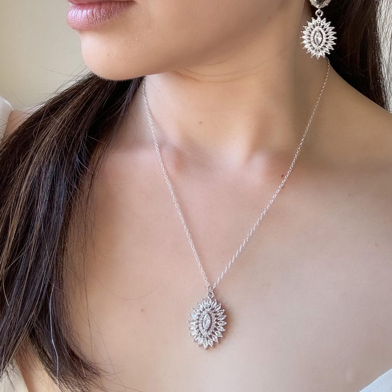 May include: A silver necklace and matching earrings set. The jewelry features a sunburst design with a central oval-shaped stone surrounded by smaller, sparkling stones. The necklace hangs on a delicate chain.