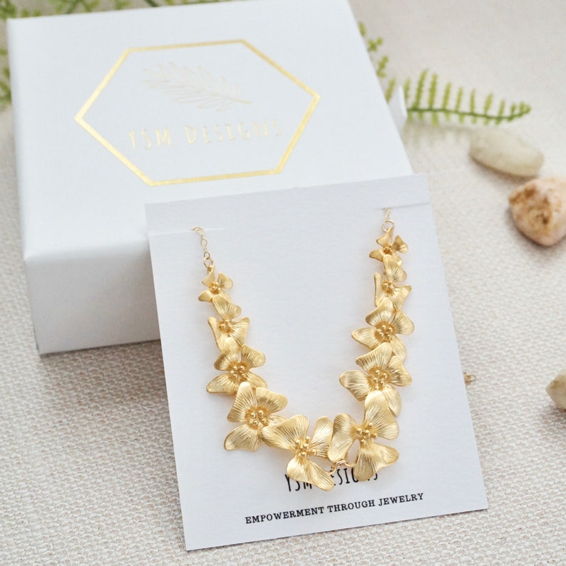 May include: Gold necklace with a delicate chain and a floral design. The necklace is displayed on a white card with the text "YSM Designs" and "Empowerment Through Jewelry".