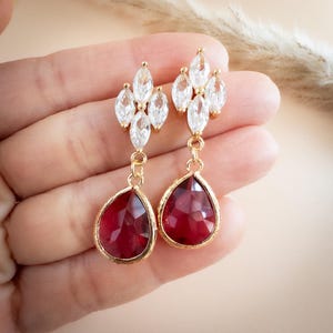 May include: Gold-plated drop earrings featuring clear crystal clusters and faceted, ruby-red teardrop gems. These earrings, designed for pierced ears, are suitable for special occasions. The earrings are a classic design, suitable for weddings or formal events.