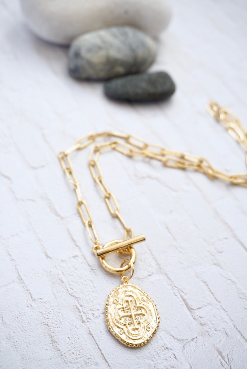 May include: A gold chain necklace with a toggle clasp and a large oval pendant featuring a cross design.