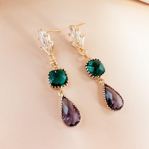 May include: A pair of gold-tone drop earrings featuring a cluster of clear baguette-cut crystals at the post, followed by a square-cut emerald green crystal and a pear-shaped purple crystal, all set in a delicate gold-tone metal frame.