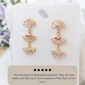 May include: Gold-toned, fan-shaped dangle earrings with clear, sparkling accents. Each earring features three graduated fans, connected by delicate chains. The earrings are displayed on a white card with text that reads, "The earrings are absolutely beautiful. They are well made and look much more expensive than their price."
