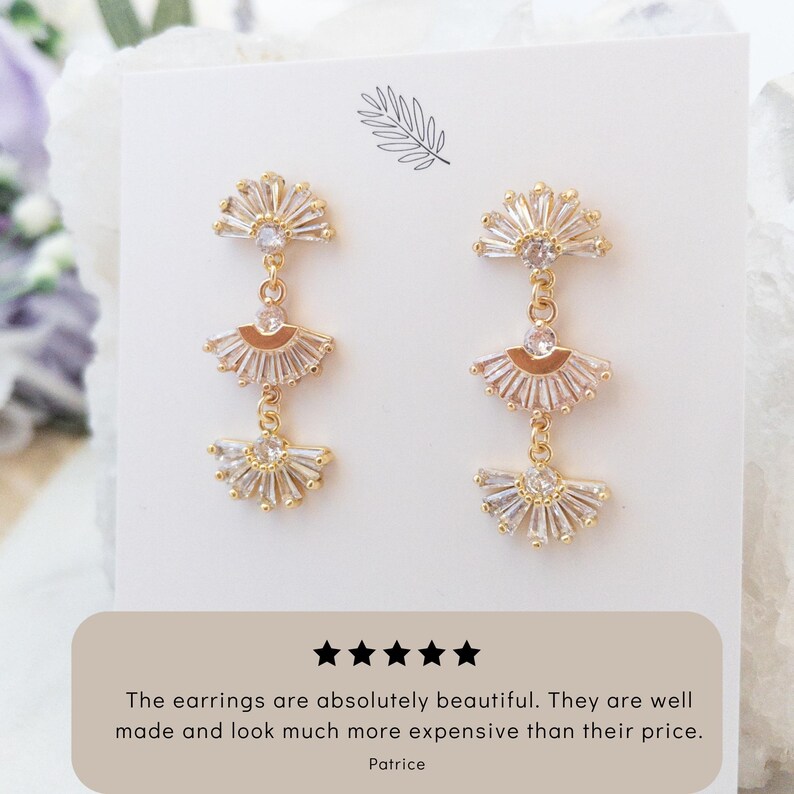 May include: Gold-toned, fan-shaped dangle earrings with clear, sparkling accents. Each earring features three graduated fans, connected by delicate chains. The earrings are displayed on a white card with text that reads, "The earrings are absolutely beautiful. They are well made and look much more expensive than their price."
