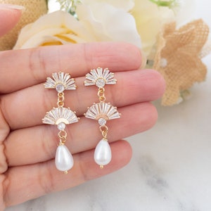 May include: Gold-toned drop earrings featuring a fan-shaped design with clear crystals and a pear-shaped white pearl. The earrings have a delicate, elegant appearance, suitable for special occasions.