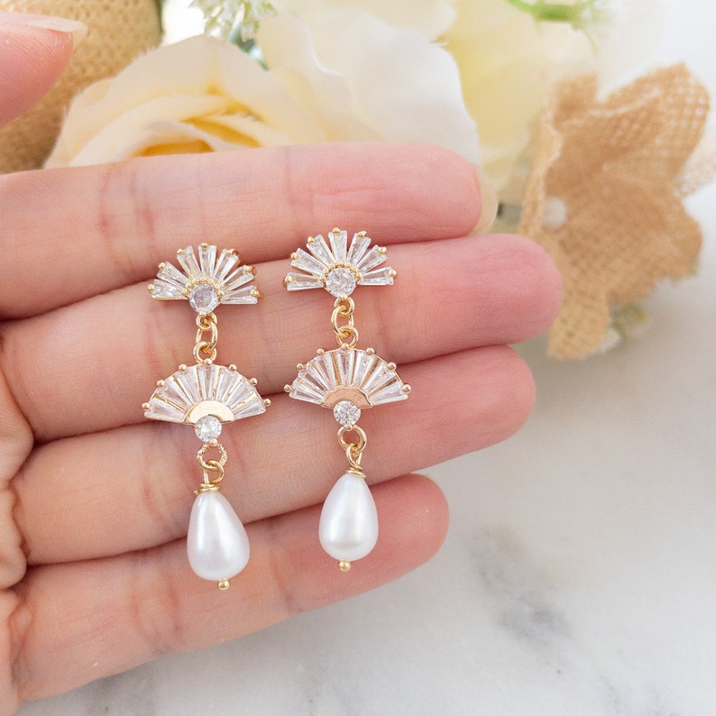 May include: Gold-toned drop earrings featuring a fan-shaped design with clear crystals and a pear-shaped white pearl. The earrings have a delicate, elegant appearance, suitable for special occasions.