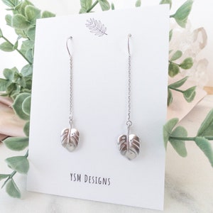 May include: A pair of silver dangle earrings with a monstera leaf charm. The earrings are on a white card with the text "YSM DESIGNS".