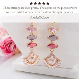 May include: Gold dangle earrings with three marquise-shaped gemstones in shades of pink, purple, and red. The earrings feature a fan-shaped design with clear cubic zirconia stones.