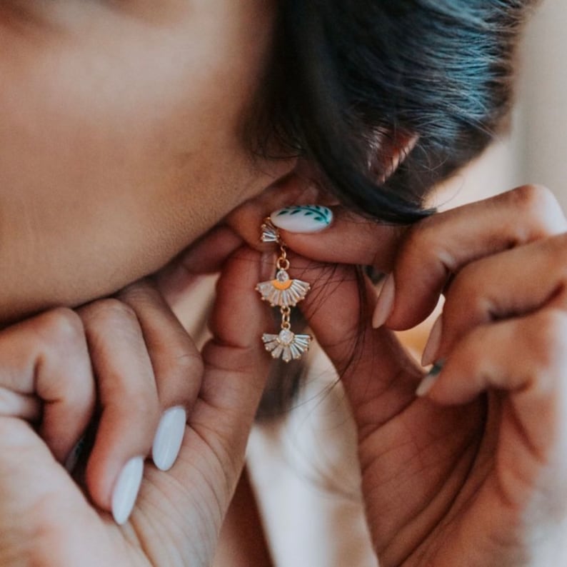 May include: Gold-toned earrings with a fan-shaped design, featuring clear, sparkling accents. The earrings are being worn, showcasing their delicate and elegant style. The design is reminiscent of Art Deco jewelry.