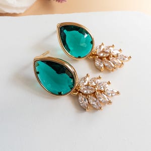 May include: A pair of gold-tone earrings featuring a large, pear-shaped emerald green gemstone set in a smooth metal frame. Attached to the base of each teardrop stone is a cluster of small, clear, marquise-cut crystals arranged in a fan shape.