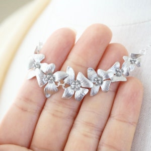 May include: A silver necklace with a delicate chain and a row of five flower-shaped pendants. The flowers are detailed with small, raised dots in the center.