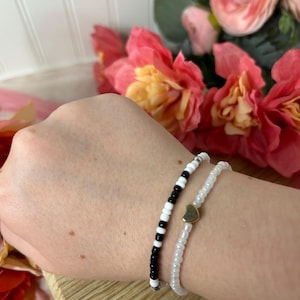 May include: Two beaded bracelets. One bracelet features black and white beads, while the other is white with a gold heart charm. The bracelets are displayed on a wooden surface, with pink flowers in the background.