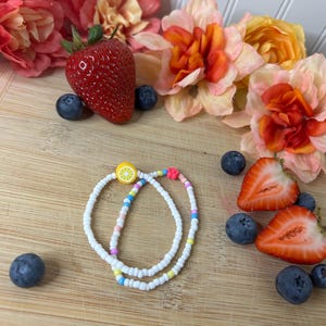 May include: Two white beaded bracelets with colorful accents, including a lemon slice and a pink heart. The bracelets are displayed with fresh strawberries, blueberries, and flowers on a wooden surface.