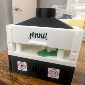 May include: A white and black box-shaped object with the name "jenna" in cursive. The box has two black doors with hazard symbols and a green object inside. The top is black and the box is on a wooden surface.