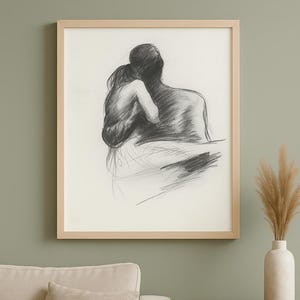May include: A framed charcoal drawing of an embracing couple. The artwork is in a light wood frame, hanging on a muted green wall. A beige sofa and a dark wood side table with a vase of dried plants are also visible.
