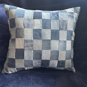 May include: Square cushion with a woven denim patchwork design in shades of blue and grey. The cushion is square and rests on a dark blue, textured surface.