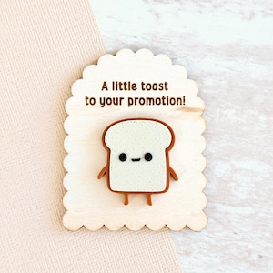 May include: A wooden plaque with a scalloped edge features a cartoon toast character with arms and legs. The text "A little toast to your promotion!" is printed above the toast.