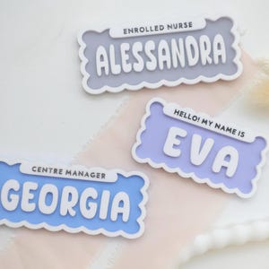 Könnte beinhalten: Drei Namensschilder mit gewellten Rändern. Das obere Schild ist grau und trägt die Aufschrift "ENROLLED NURSE" über dem Namen "ALESSANDRA". Das mittlere Schild ist lavendelfarben und trägt die Aufschrift "HELLO! MY NAME IS" über dem Namen "EVA". Das untere Schild ist blau und trägt die Aufschrift "CENTRE MANAGER" über dem Namen "GEORGIA".