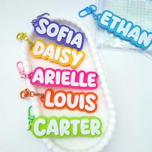 May include: A selection of colourful name keychains, each with a different name in white text on a coloured background. The keychains include names like Sofia, Daisy, Arielle, Louis, and Carter. A blue keychain with the name Ethan is also visible.