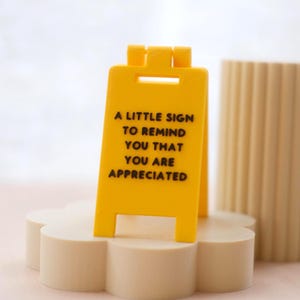 May include: A bright yellow sign with black text: "A LITTLE SIGN TO REMIND YOU THAT YOU ARE APPRECIATED." The sign stands on a beige, flower-shaped base. A beige, ribbed cylindrical object is in the background.