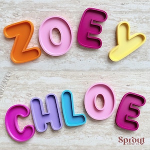 Op de afbeelding: Kleurrijke letters in de vorm van koekjessnijders die de namen "ZOEY" en "CHLOE" spellen. De letters zijn in verschillende kleuren: oranje, roze, magenta en geel voor "ZOEY", en magenta, paars, blauw, roze en magenta voor "CHLOE". Het logo "Sprout Paper Co." staat rechtsonder.