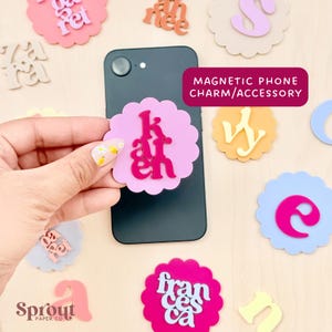 May include: A black smartphone with a pink scalloped magnetic phone charm that says "Karen" in a bold font. The charm is attached to the phone. Other colorful name charms and the text "MAGNETIC PHONE CHARM/ACCESSORY" are also visible.