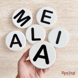 May include: White, circular coasters with black letters spelling "MEALIA". One coaster is held in a hand. The coasters are on a light-coloured surface. The Sprout Paper Co. logo is in the bottom right corner.